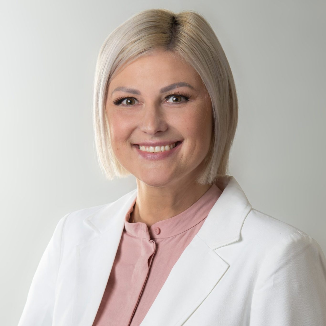 Portrait Zahnärztin Anna Mann Portrait Zahnärztin Anna Mann: Frau mit blonden Haaren, blassem Teint und in weißer Blazerjacke, lächelnd.